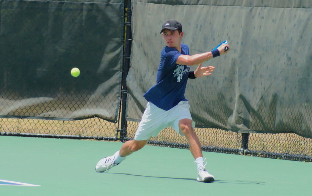 A tennis player in a blue shirt and white shorts prepares to hit a forehand shot on an outdoor court.