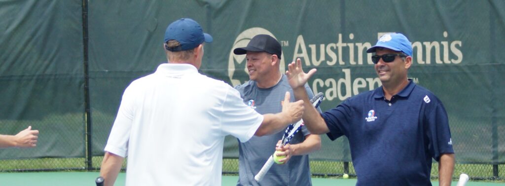 Three men on a tennis court high-five and smile, with Austin Tennis Academy banner in background.