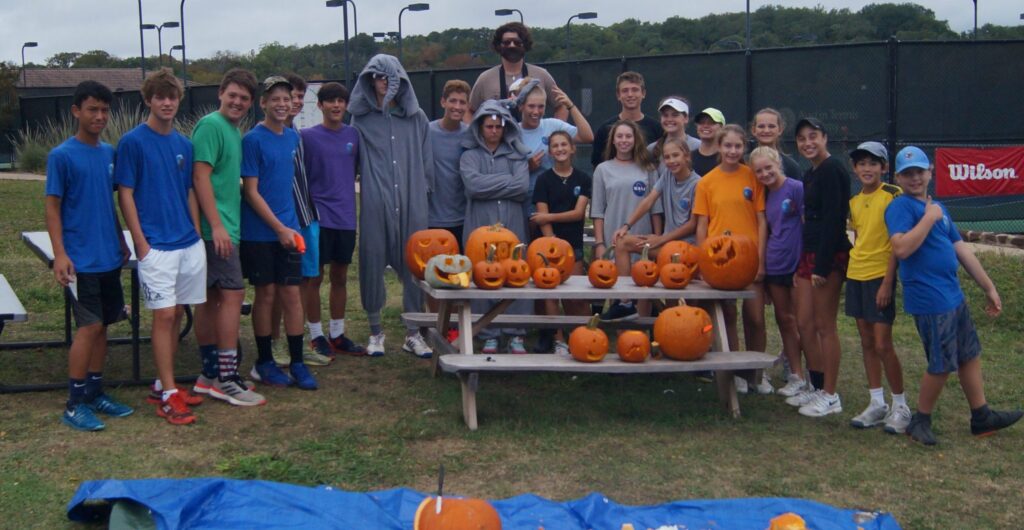 A group of teens and kids posing outdoors with carved pumpkins on picnic tables at a tennis court.