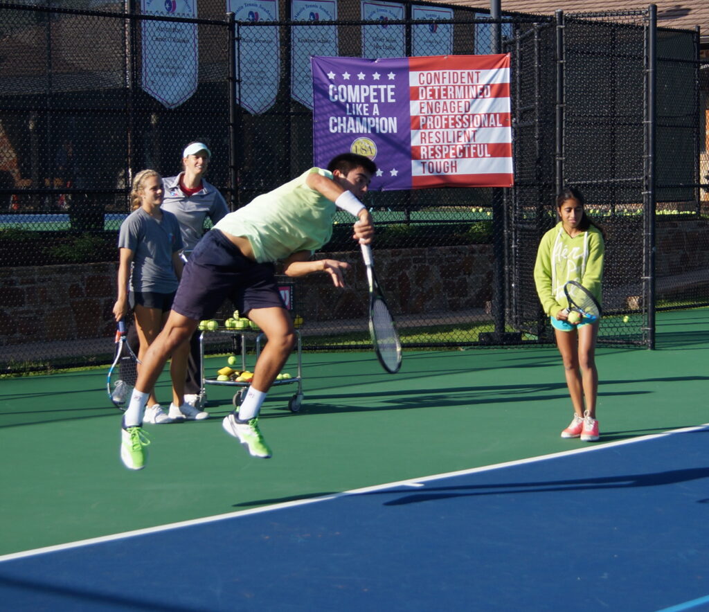 A tennis player jumps to hit a shot while others watch on an outdoor court during practice.