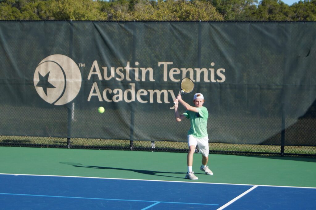 A tennis player in a green shirt hits a backhand shot on an outdoor court at Austin Tennis Academy.