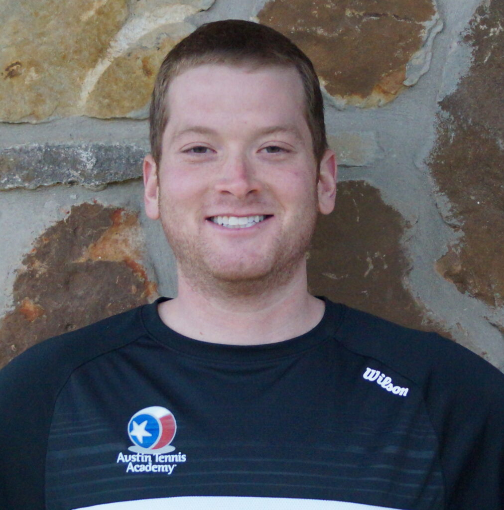 Smiling man in a black Austin Tennis Academy shirt standing in front of a stone wall.