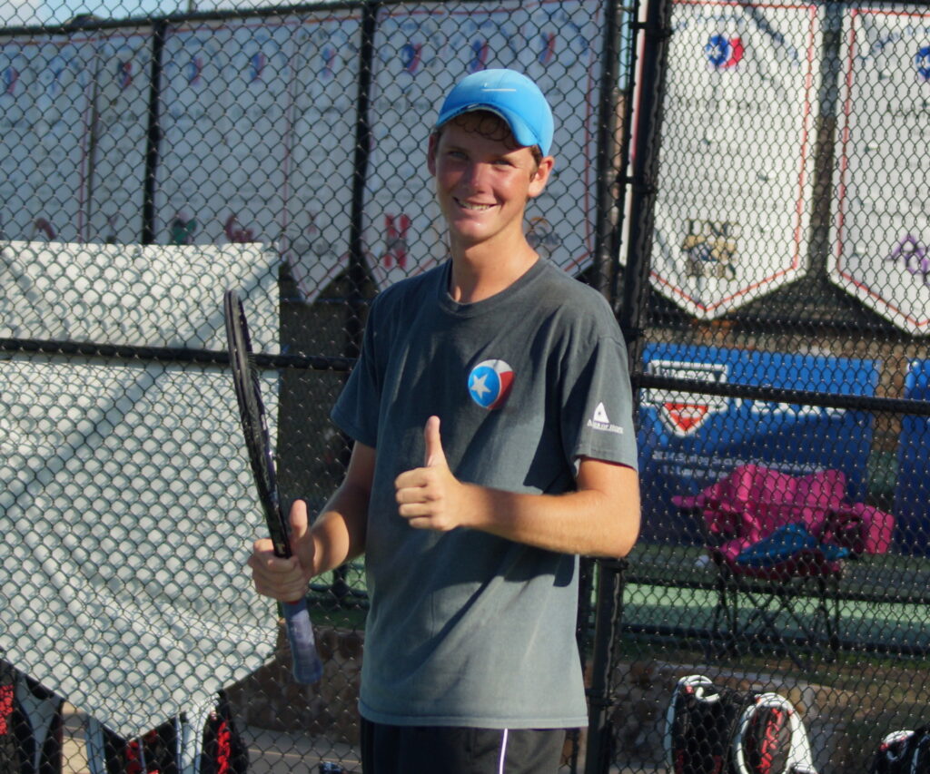 Teen tennis player in a blue cap smiles and gives a thumbs up while holding a racquet near a court fence.