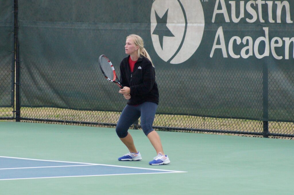 A woman holding a tennis racquet stands ready on an outdoor court near a fence with Austin Academy on it.