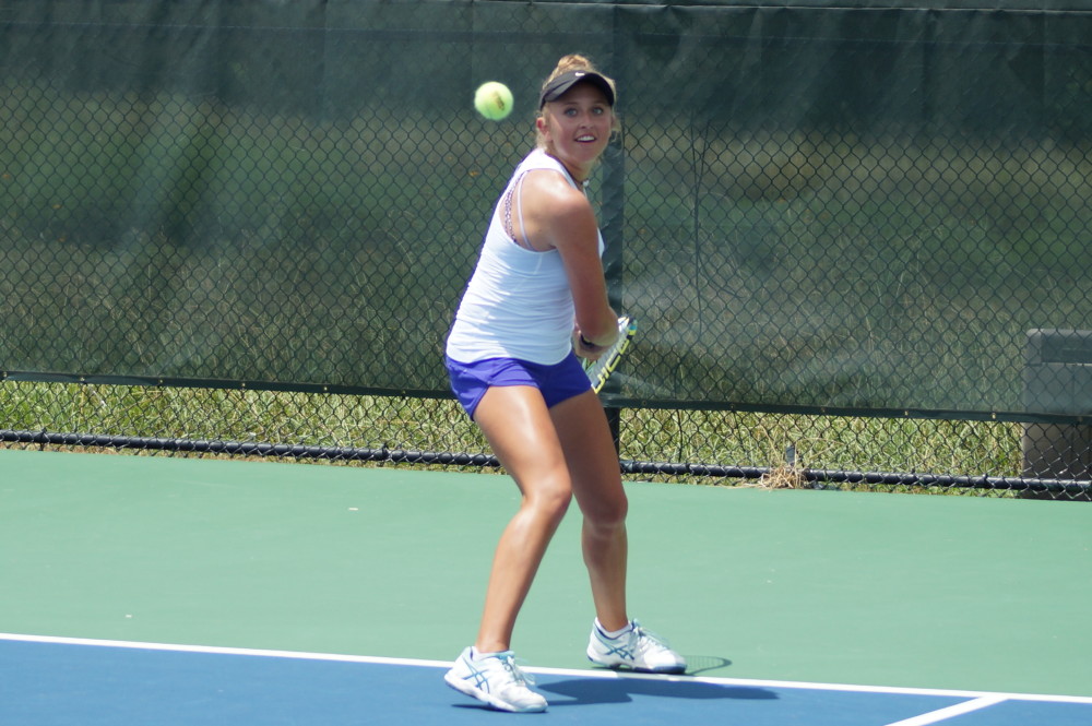 Woman in tennis attire preparing to hit a backhand shot on an outdoor court with a fence in the background.