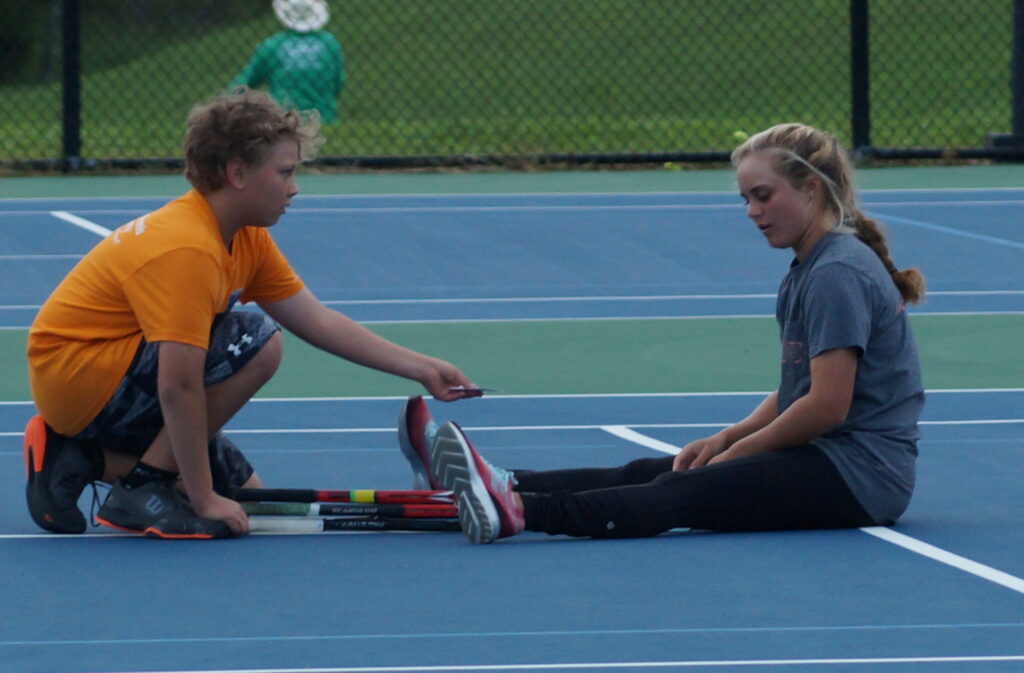 Two kids sit on a tennis court, one handing something to the other, with rackets lying on the ground.