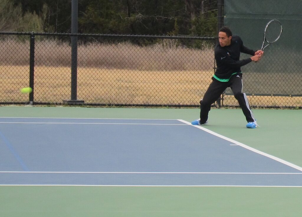 A tennis player prepares to hit a backhand shot on an outdoor court, with the ball approaching.