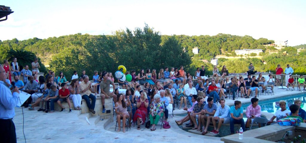 A large group of people sit outdoors by a pool, listening to a speaker, with trees in the background.