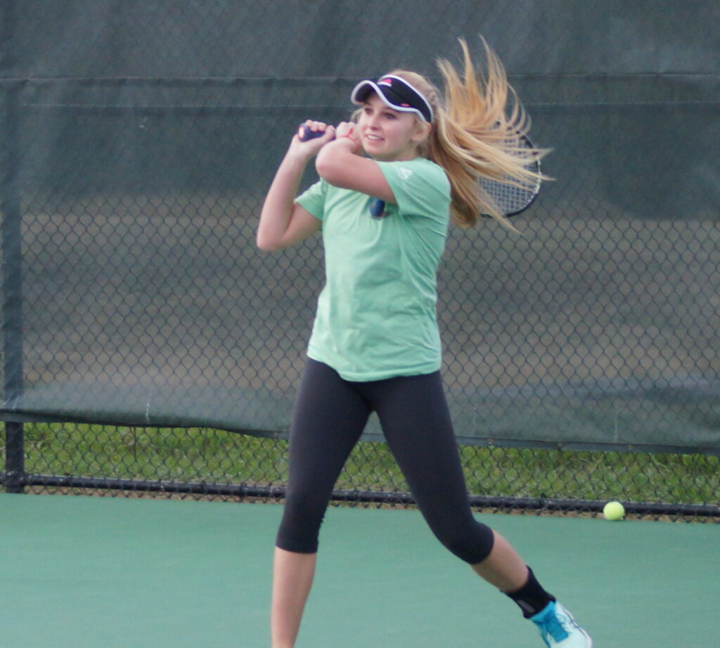 A young woman swings a tennis racket on an outdoor court, wearing athletic clothes and a visor.