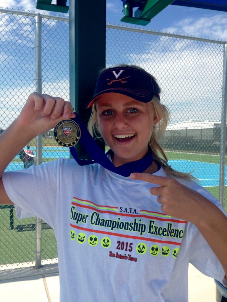 Smiling girl in a tennis shirt points to a medal, standing by blue tennis courts on a sunny day.