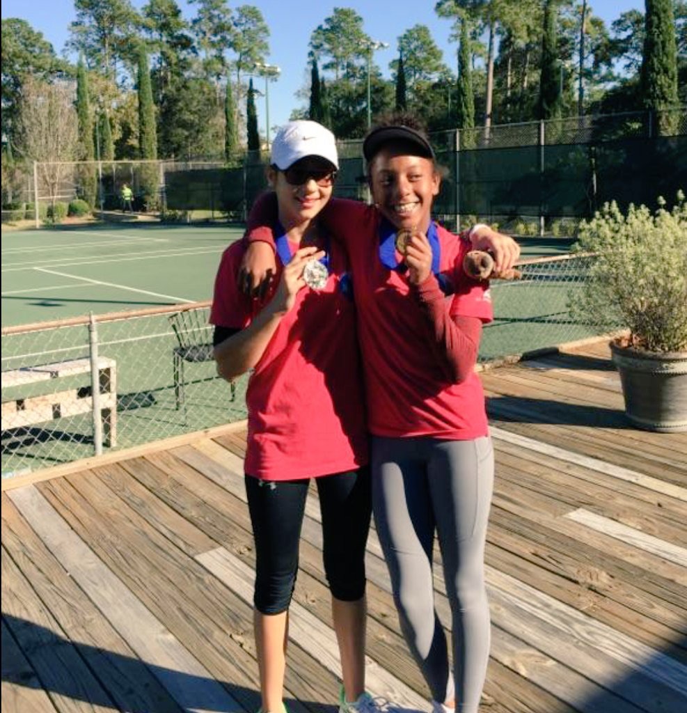 Two girls in red shirts on a tennis court, smiling and showing medals with arms around each other.