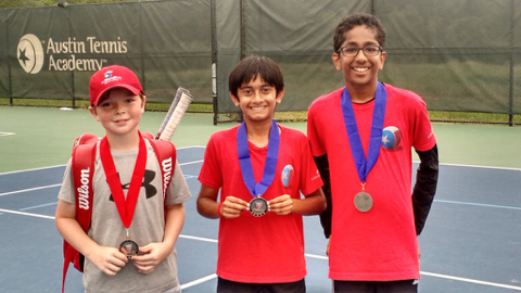 Three boys with medals stand on a tennis court, smiling, in front of an Austin Tennis Academy sign.