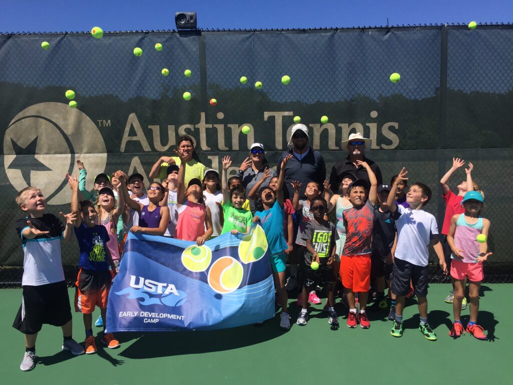 Group of kids and coaches on a tennis court, tossing tennis balls in the air, holding a USTA camp banner.
