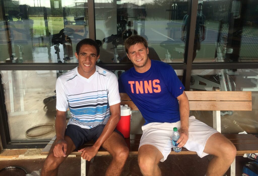 Two men in athletic wear sit on a bench by a gym window, smiling after playing tennis.