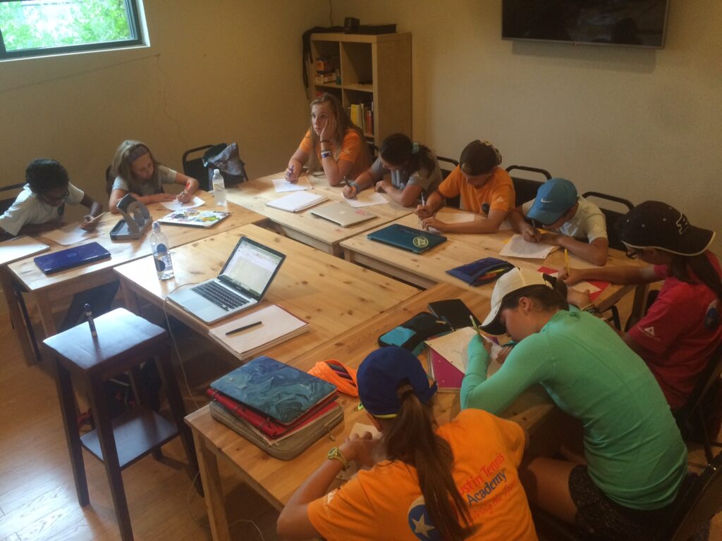 Eight students and a teacher sit at tables in a classroom, writing and working on assignments.
