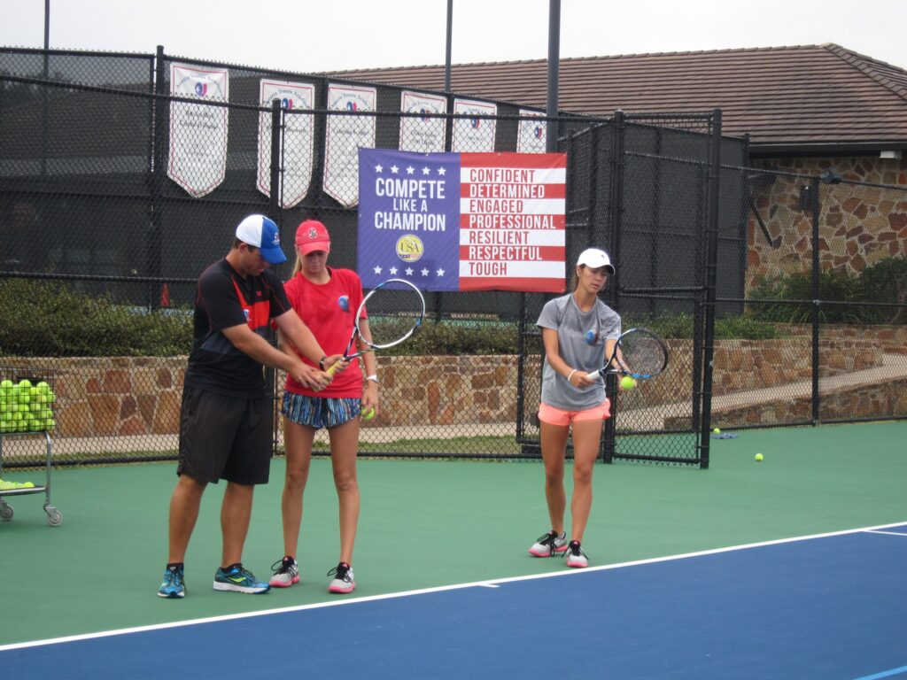 A tennis coach instructs two young women with rackets on an outdoor tennis court near a motivational sign.