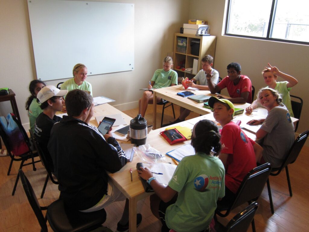 A group of students sit around tables in a classroom, working on assignments and smiling at the camera.