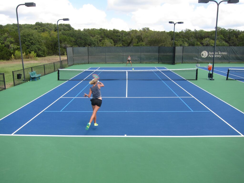 Two women playing tennis on an outdoor blue and green court under a partly cloudy sky.