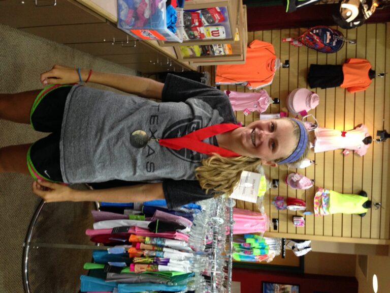 Smiling girl wearing a medal stands in a clothing store with colorful shirts and accessories on display.