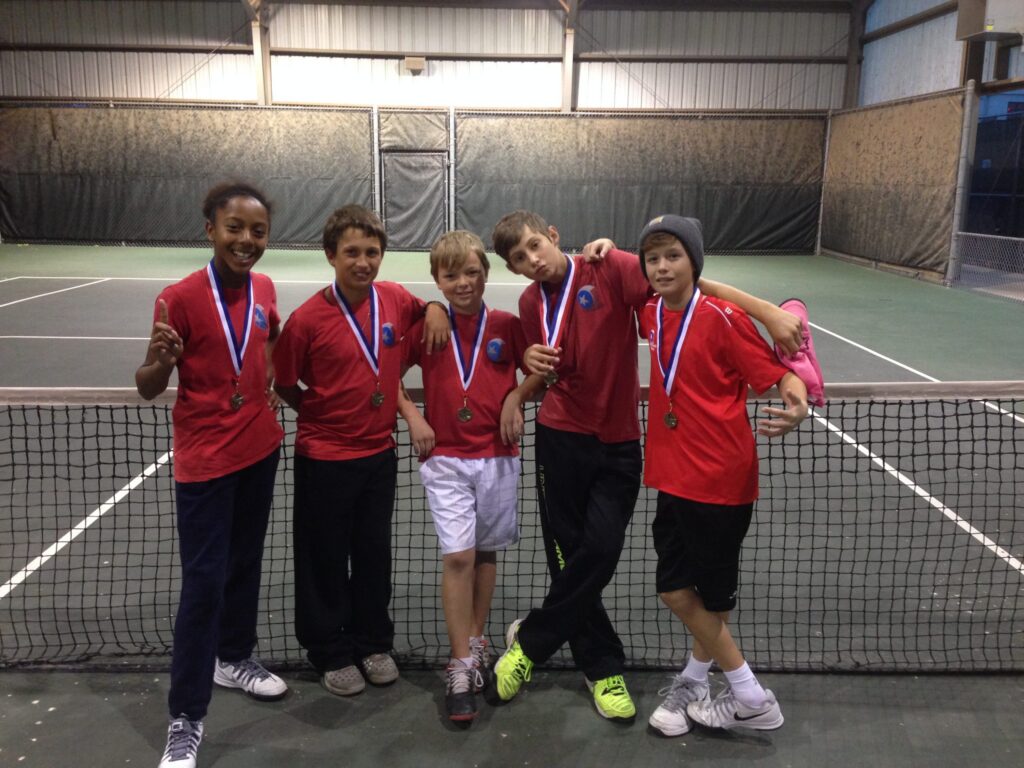 Five kids in red shirts with medals pose together, smiling, on an indoor tennis court in front of the net.