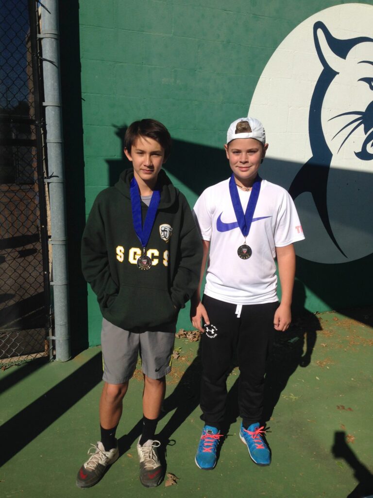 Two boys wearing medals stand on a tennis court in sunlight, with a wildcat logo on the green wall behind them.