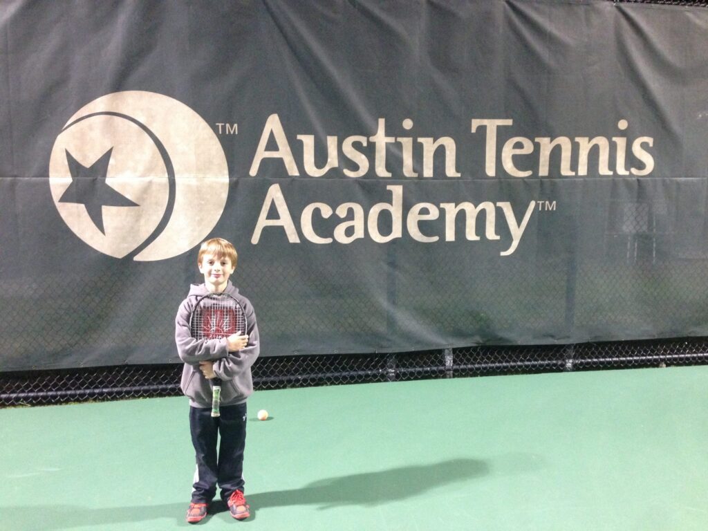 Young boy holding a tennis racket stands on a court in front of an Austin Tennis Academy banner.