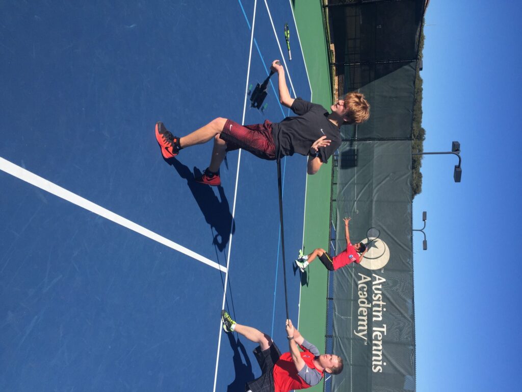 Three people play tennis on blue courts at Austin Tennis Academy, all mid-air with rackets raised.