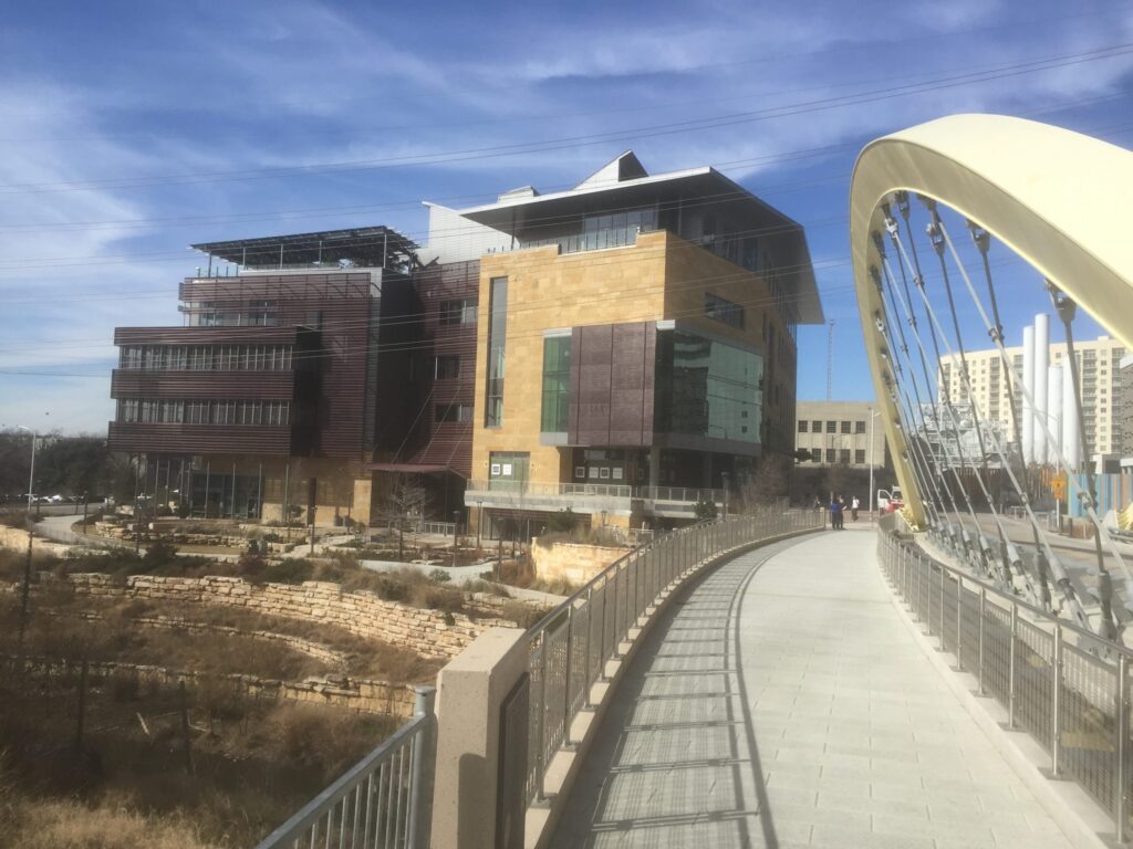 Modern multi-story building next to a pedestrian bridge under a partly cloudy blue sky.