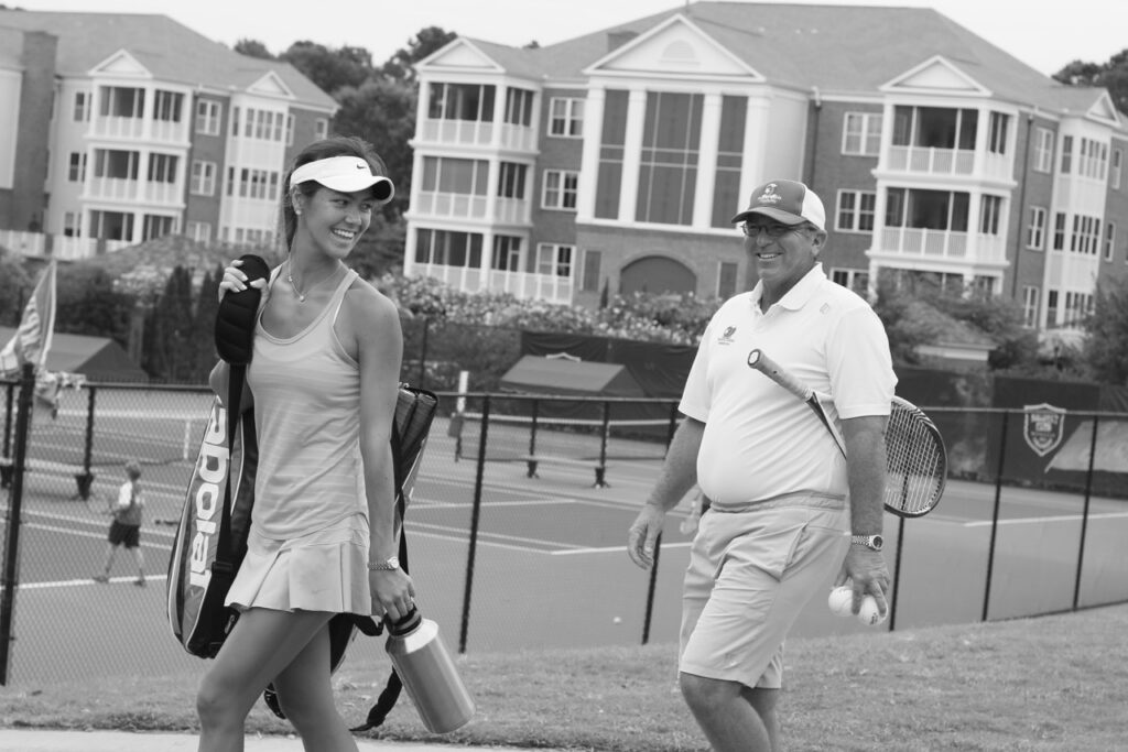 Woman and man with tennis gear walk by tennis courts, smiling, with buildings in the background.
