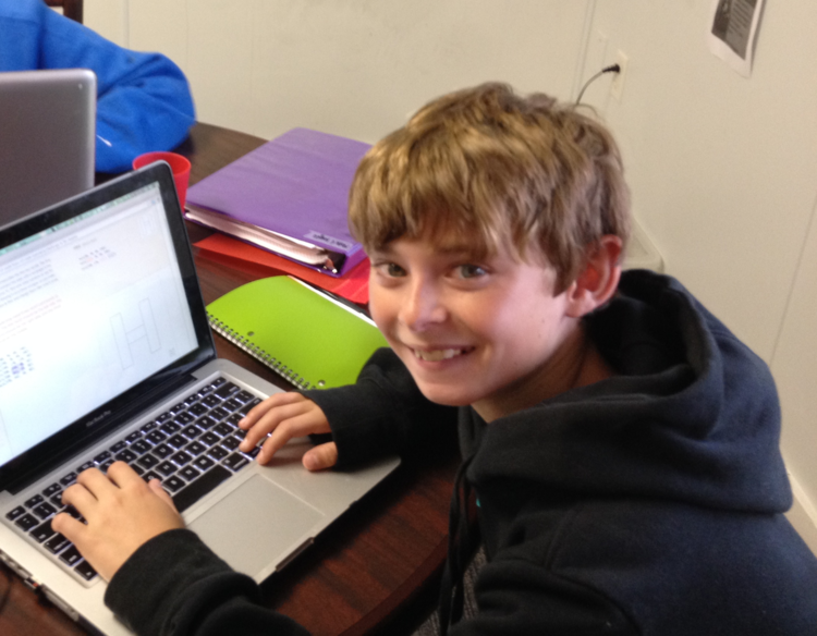 Smiling boy in a black hoodie using a laptop at a desk with notebooks and folders nearby.
