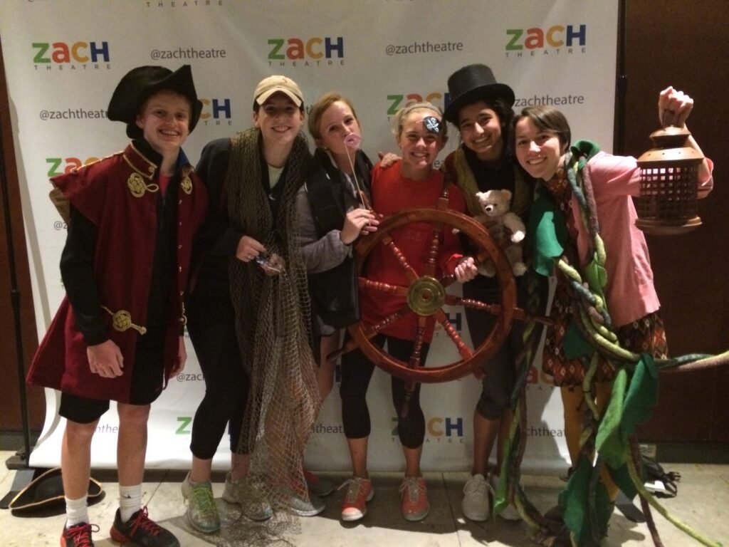 Six people in playful costumes pose together, smiling in front of a ZACH Theatre step-and-repeat banner.