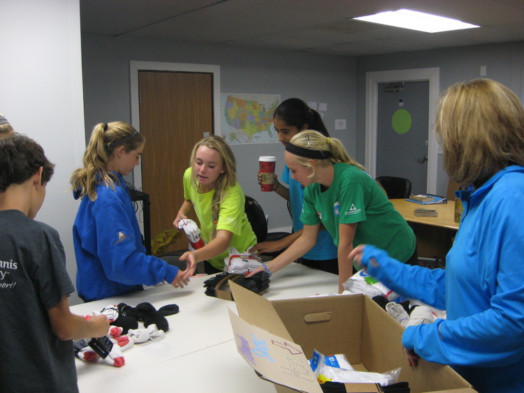 A group of teens and an adult sort and pack socks in a room with tables and boxes.