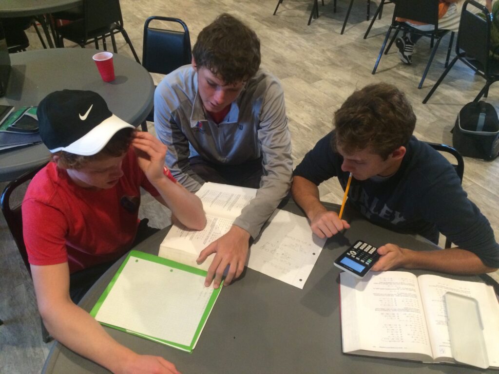 Three boys sit at a table working on math homework with papers, a calculator, and textbooks.