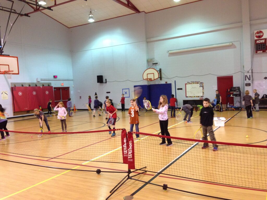 Children in a gymnasium play badminton, holding rackets and shuttlecocks near a net during a PE class.