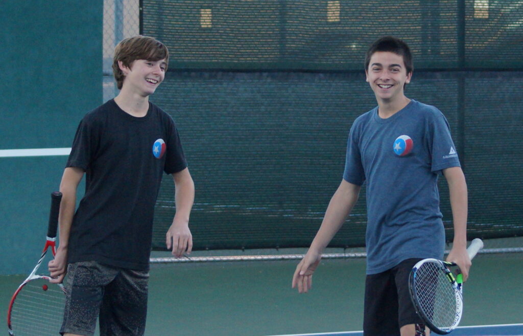Two teenage boys smiling and holding tennis rackets on an outdoor tennis court.