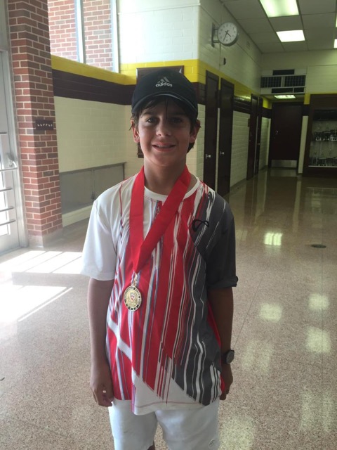 A boy wearing a medal and striped shirt stands in a brightly lit school hallway, smiling at the camera.