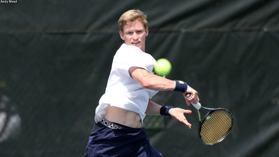 A tennis player in a white shirt swings a racket to hit a green tennis ball on an outdoor court.