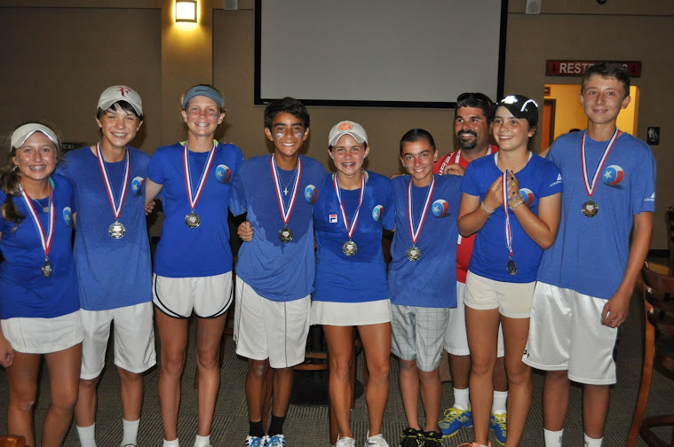 Eight smiling young tennis players in blue shirts and medals pose indoors with their coach.