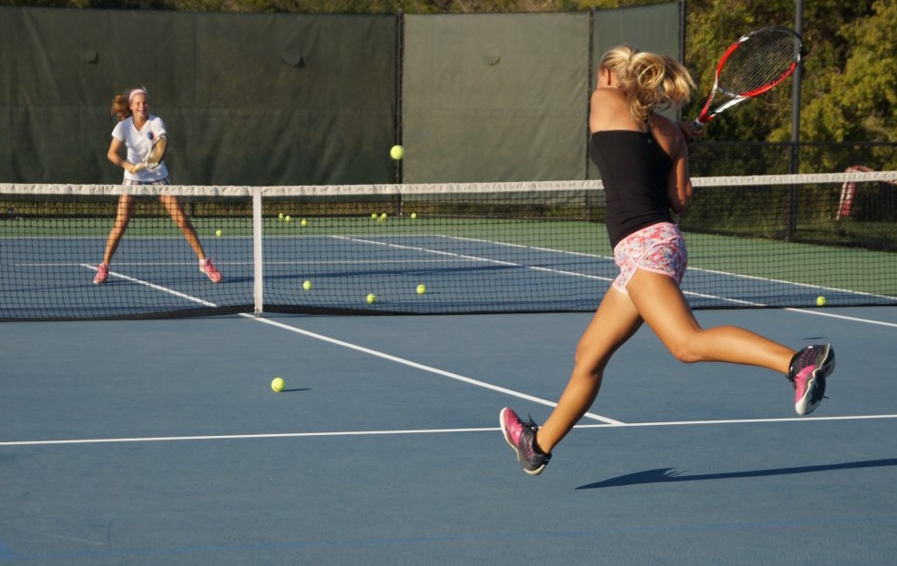 Two women playing tennis on an outdoor court, with tennis balls scattered across the surface.