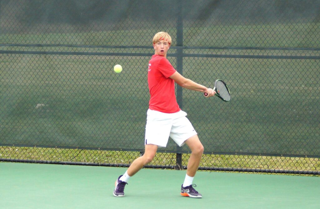 A tennis player in a red shirt and white shorts prepares to hit a backhand shot on an outdoor court.