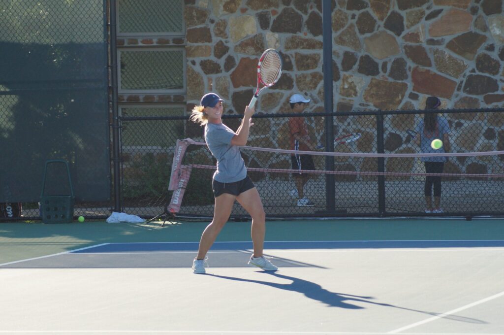 Person in athletic wear hitting a tennis ball with a racket on an outdoor court; others visible in background.