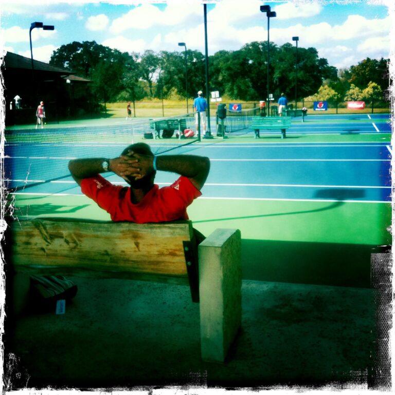 A person in a red shirt relaxes on a bench by outdoor tennis courts on a sunny day.