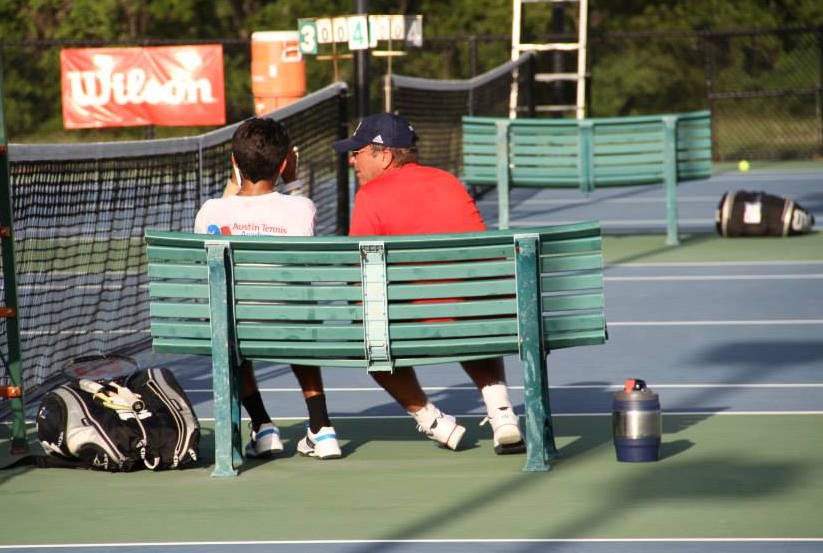 Two people sitting on a bench by a tennis court, talking, with tennis gear and a water jug nearby.