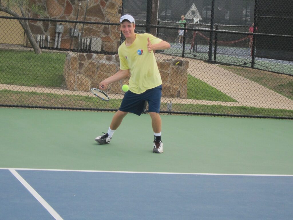 Man on a tennis court smiling, giving a thumbs up, holding a racket, and preparing to hit a tennis ball.