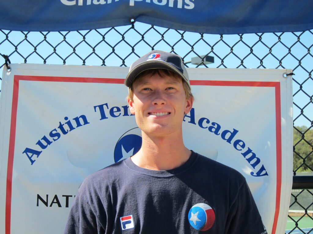 Young man in a cap and T-shirt stands smiling in front of an Austin Tennis Academy banner outdoors.