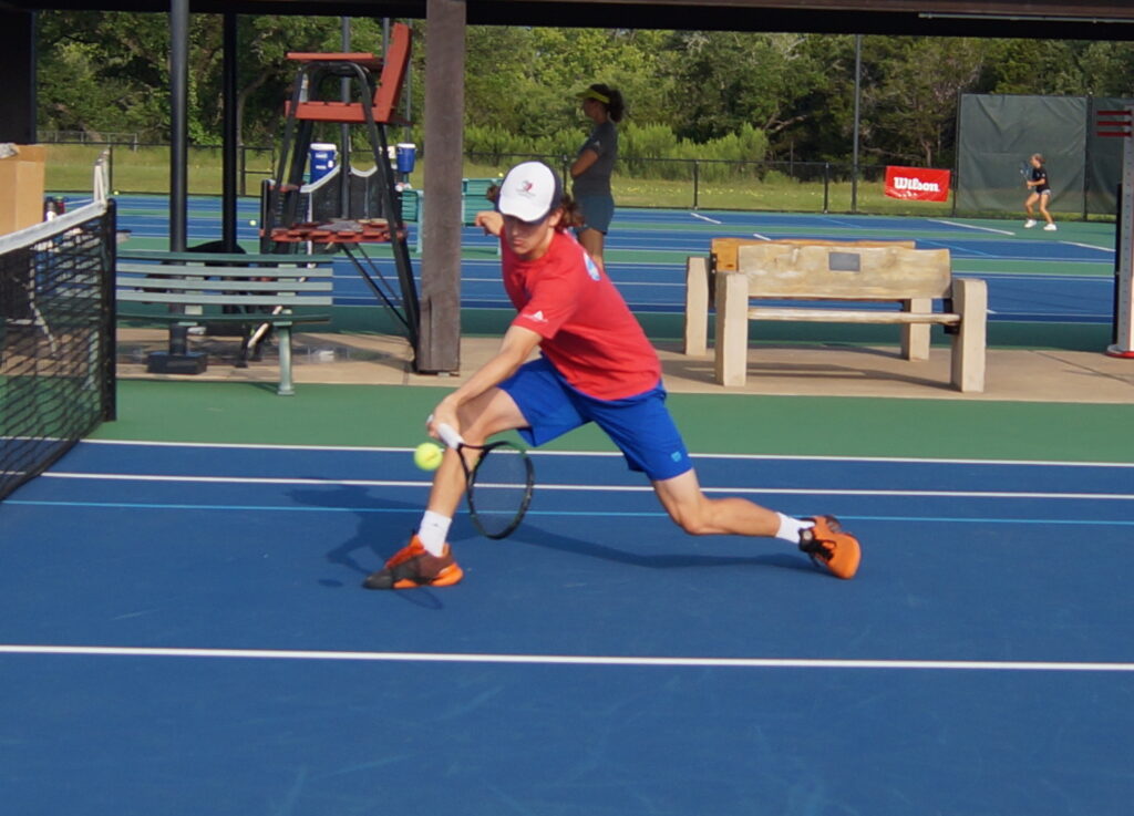 Tennis player in red shirt and blue shorts reaching to hit a low shot on an outdoor court.