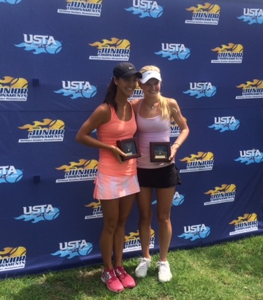 Two young women holding awards pose in front of a blue USTA Junior Tournament backdrop on grass.