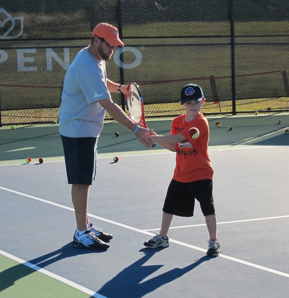 An adult teaches a young boy how to hold a tennis racket on an outdoor court.