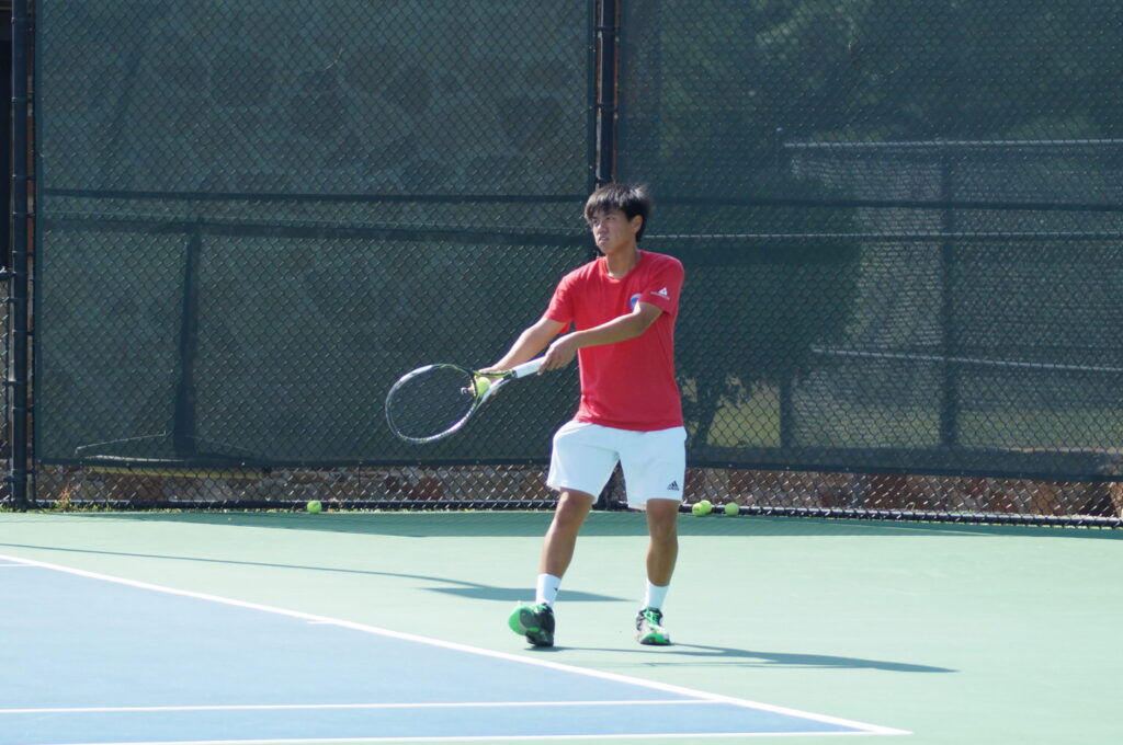 A tennis player in a red shirt prepares to hit a forehand shot on an outdoor tennis court.