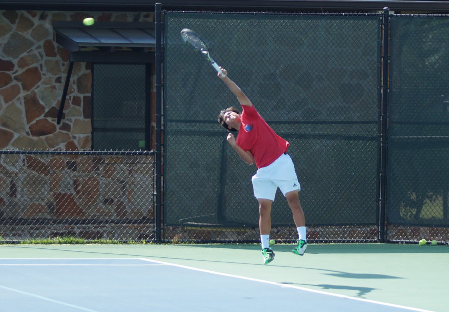 A tennis player in a red shirt jumps to serve the ball on an outdoor court.